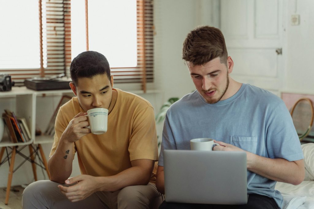 Two men at home with coffee and laptop, sharing a casual moment.